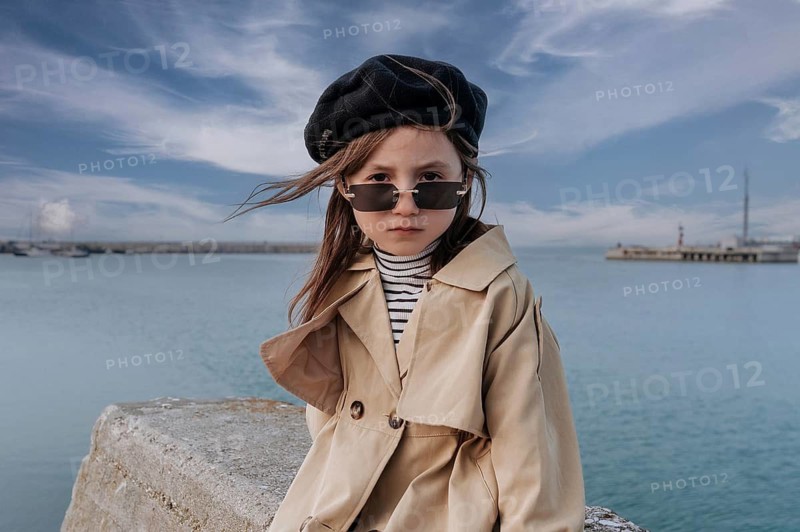 A young girl in trendy attire stands by the waterfront in Civitanova, Italy. Her stylish trench coat, beret, and sunglasses add flair as she enjoys the breezy seaside view