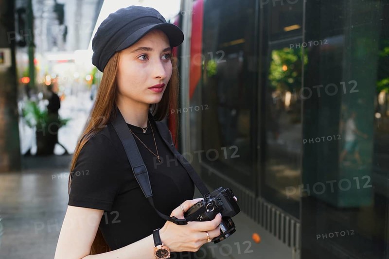 A young woman photographer in a black cap holds a camera, focusing intently on capturing city life. The urban setting is bustling with activity, reflecting vibrant city energy