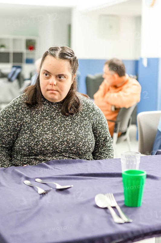 A woman sits at a table in a mental health residence, appearing deep in thought. The setting is a dining area with utensils and a green cup on the table