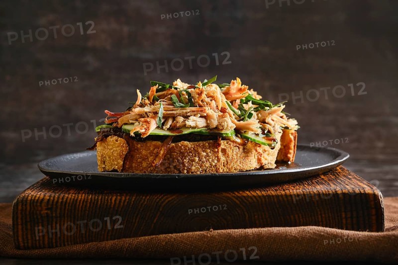 Pulled pork on top of a slice of toast with caramelized onions, cucumber and barbecue sauce. The dish is presented on a dark plate placed on a brown cloth, showing a casual dining setup