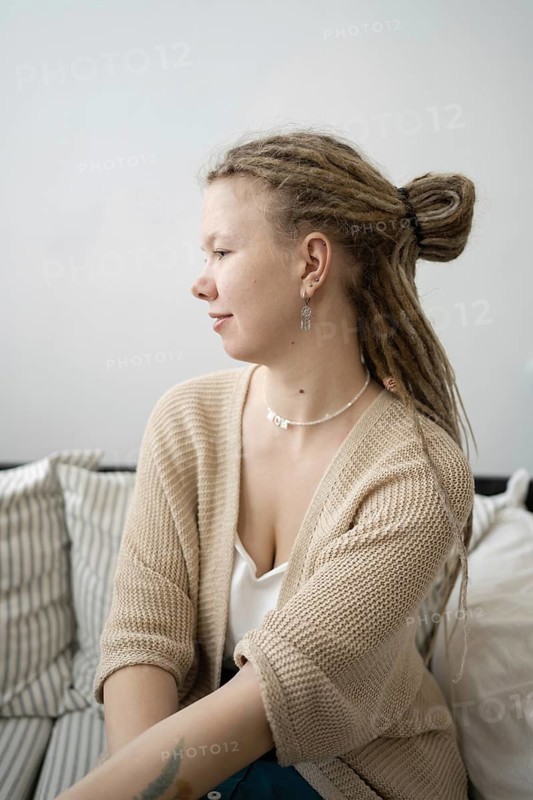 A young woman with dreadlocks and stylish earrings sits on a comfortable sofa She is wearing a beige cardigan and a white top, creating a relaxed and cozy atmosphere