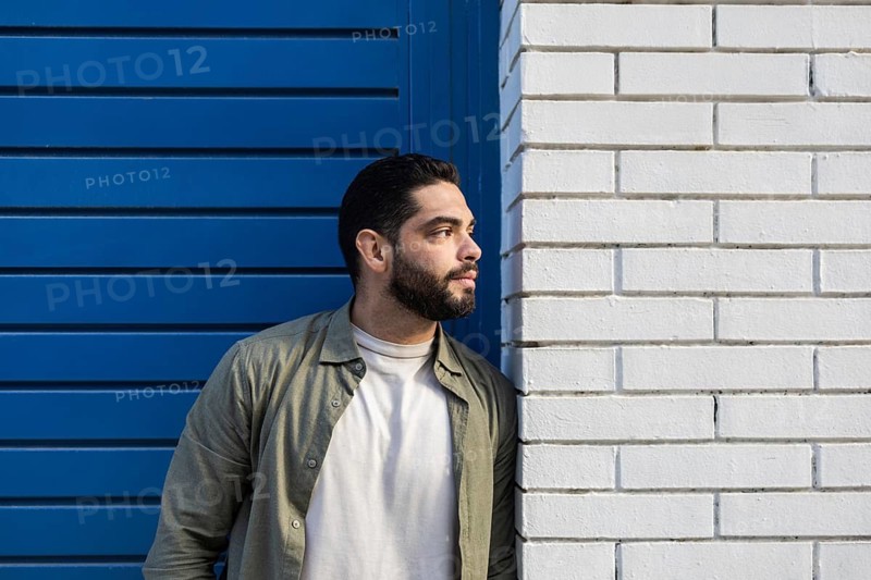 A man in a casual shirt leans against a white brick wall of the city, standing next to a blue shutter. He gazes thoughtfully into the distance, suggesting contemplation