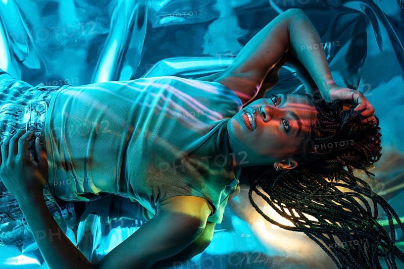 A futuristic African young woman lies surrounded by neon lights in a vibrant metaverse environment. Her braided hairstyle and innovative atmosphere create a striking visual