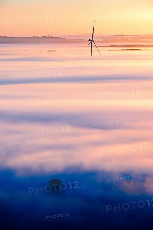 View of a scenic landscape in morning fog at sunrise with a wind turbine sticking out of the fog