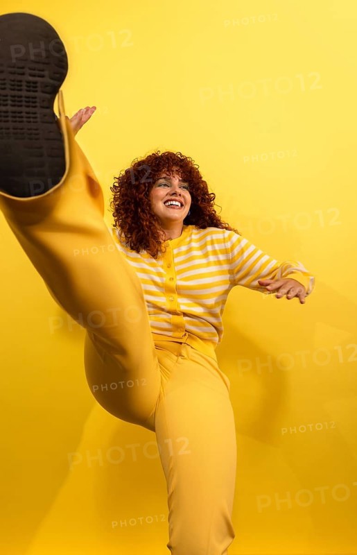 Cheerful woman with curly hair in a yellow striped sweater and pants, joyfully kicking in the air against a vibrant yellow background, embodying fun and energy