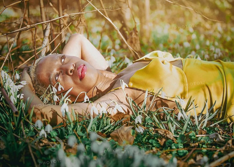 A woman in a golden dress lies peacefully among blooming snowdrops, basking in sunlight in Tacony Creek Park. The tranquil setting evokes a sense of calm and connection with nature