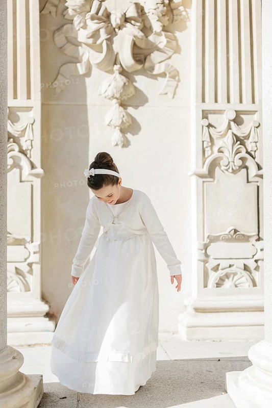 A young girl elegantly dressed in a First Communion dress stands amidst ornate architectural details, bathed in gentle sunlight, exuding a serene and holy ambiance