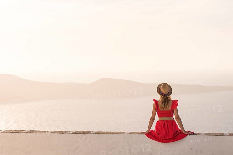 A woman in a red dress sits on a white wall, facing the tranquil seascape in Plaka, Milos island, Greece. She wears a hat, capturing a peaceful moment at sunset with distant hills visible