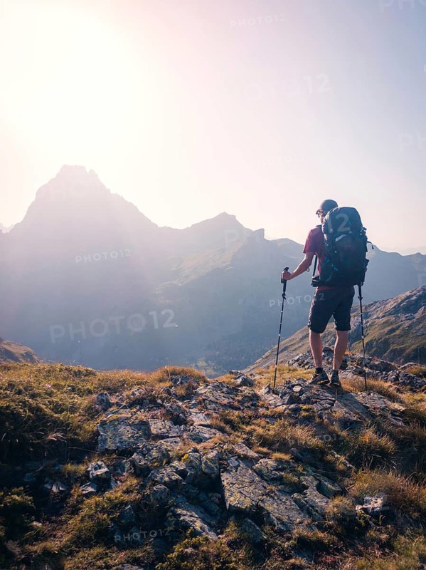 A person stands atop a rocky mountain path at sunrise, holding trekking poles in the Pyrenees. The warm sunlight casts shadows on the rugged terrain, creating an inspiring adventure scene in summer