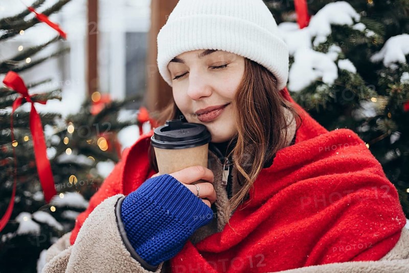 A young woman wrapped in a red scarf sips coffee near a Christmas tree on a snowy day, a moment of warmth and comfort