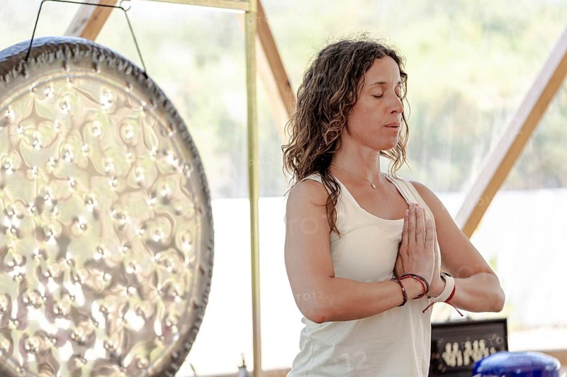 A female sound therapist meditates with a large gong in the background, practicing sound therapy. She is engaged in a holistic wellness retreat, focusing on relaxation