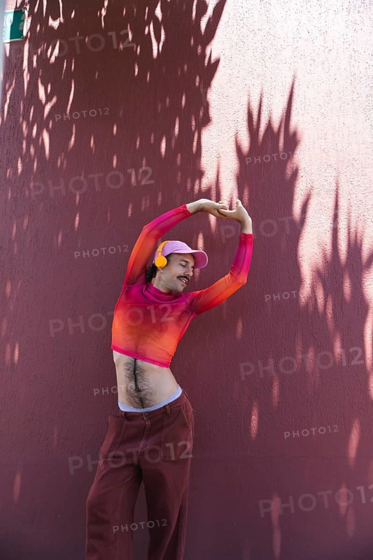 A Spanish man with a mustache, dressed in vibrant pink attire, is captured joyfully dancing against a wall in Quito, Ecuador. The cheerful expression, suggesting themes of self-expression and diversity related to the LGTBIQ+ community
