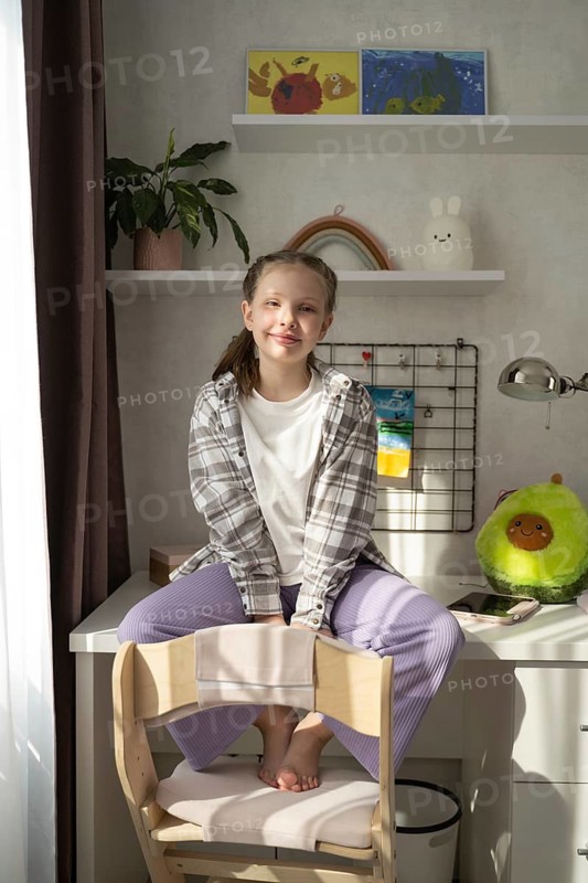 A little girl sits barefoot in her well-lit bedroom, smiling softly as she rests on a white desk. Surrounded by cheerful decor and warm sunlight, the atmosphere is calm and inviting