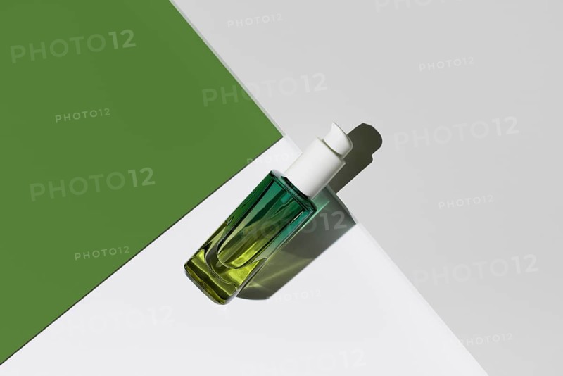 Top view of a green glass bottle with a dropper, filled with essential oil, sits on a clean white stand against a contrasting green background, showcasing a minimalist setup ideal for natural skincare