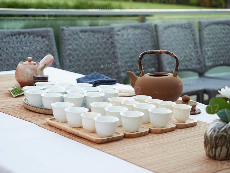 Elegant tea service served on outdoor table. Terracotta teapot, white cups on wooden trays on bamboo mat
