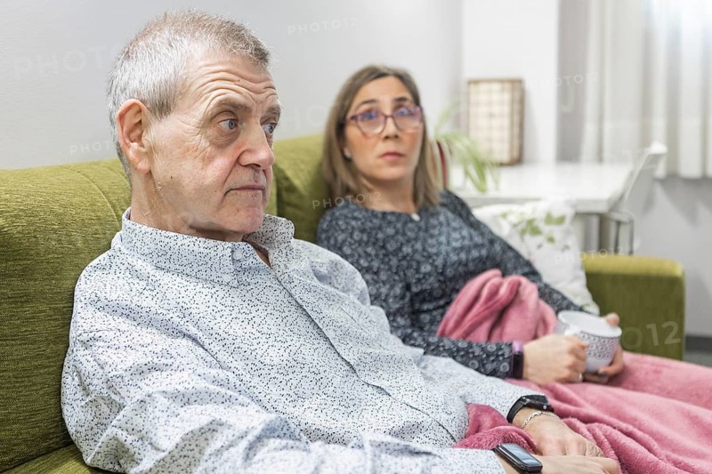 Couple with intellectual disabilities sitting on a green couch at supervised home, wrapped in a pink blanket. They are relaxed, with a cup of coffee, enjoying leisure time