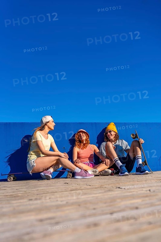 Three young skateboarders are sitting on a wooden surface against a blue wall, enjoying a sunny day and chatting