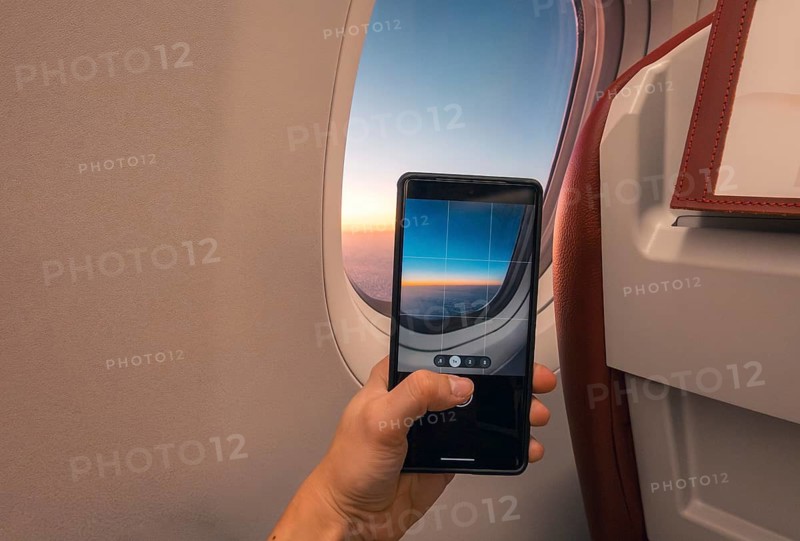 Cropped unrecognizable person captures a sunset over the ocean using a smartphone, viewed through an airplane window