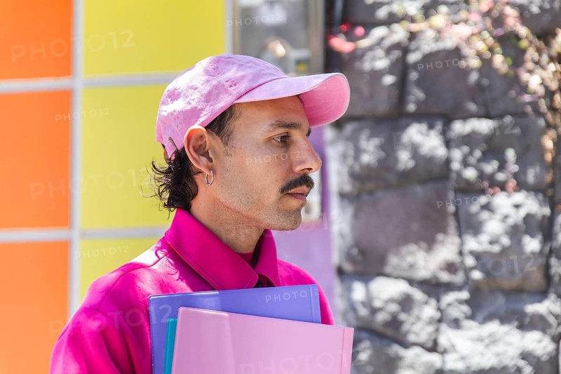 A young Spanish man with a mustache, dressed in vibrant pink, captured while experiencing the city life. This image highlights urban lifestyle, casual fashion, and expressions of joy and diversity, resonating with LGTBIQ+ themes