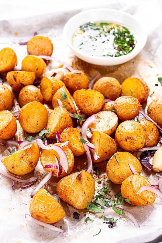 Roasted potatoes sit on a baking sheet next to chopped herbs and red onions. They are golden brown and ready to be served. Ingredients add color and flavor to the dish