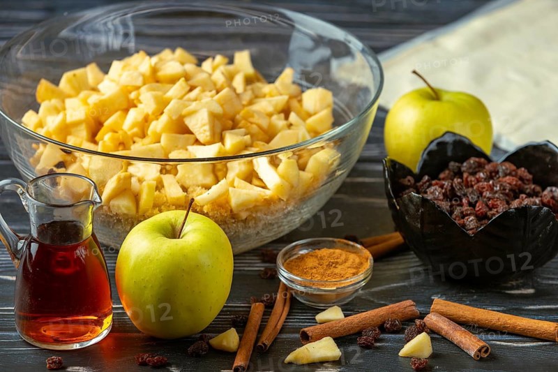 A bowl of sliced apples and baking ingredients such as raisins, cinnamon, and rum on a wooden table