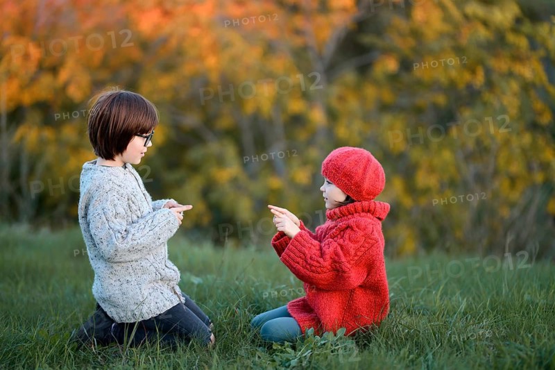 Siblings, enjoy a playful day outdoors in a park surrounded by autumn colors. The boy wears a cozy sweater and glasses, while the girl is dressed in a vibrant red outfit with a matching hat, highlighting the seasonal atmosphere