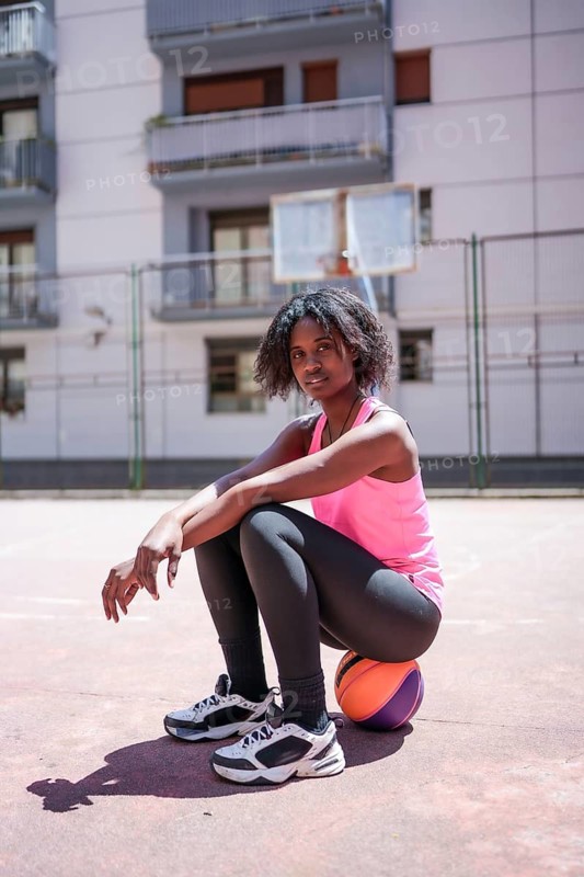 Vertical portrait of an African american woman looking at camera sitting on basketball ball outdoors