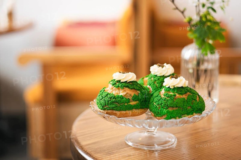 Three green matcha choux pastries topped with whipped cream served on a glass platter of the cafe. The wooden table and soft focus background add a cozy, inviting atmosphere