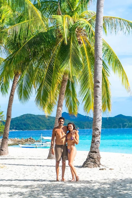Couple embracing on beautiful white sand beach with turquoise water and palm trees on malcapuya island, coron, philippines