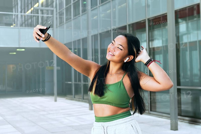 A joyful teenager takes a selfie in a modern urban setting, wearing a green top and wireless earbuds. The scene conveys a sense of youth, fun, and technology