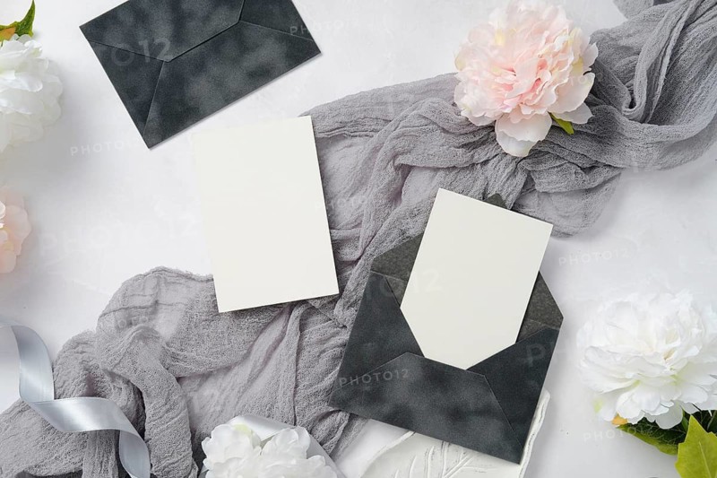 Elegant wedding stationery set with dark envelopes and blank cards. Accompanied by soft gray fabric and white peony flowers, creating a sophisticated and minimalist look