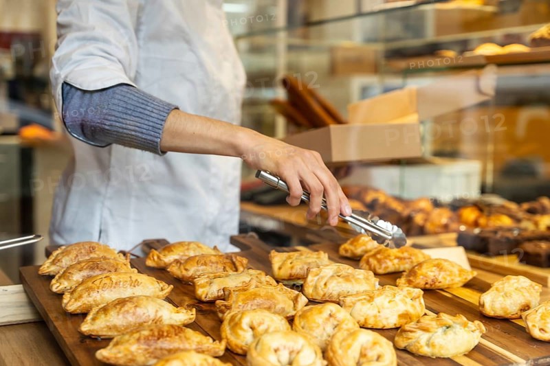 Baker's hand using tongs to select a freshly baked savory empanada from a wooden tray displayed in a glass bakery case, offering delicious food and service