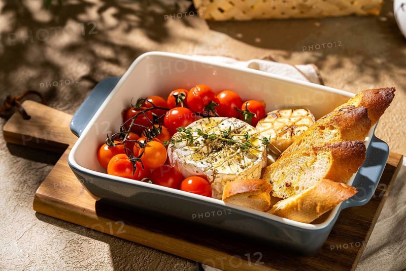 Savory baked Camembert cheese, garnished with fresh herbs, surrounded by cherry tomatoes and roasted garlic, served with crispy baguette slices in a sunlit baking dish scene