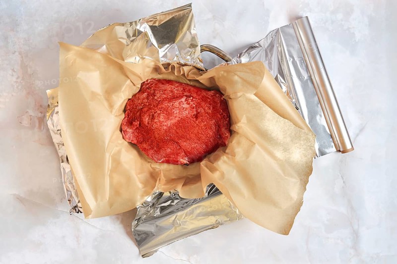 Pork shoulder is placed in a baking pan wrapped in parchment paper and aluminum foil. The setup shows the meat ready for roasting in oven. Slow cooking technique