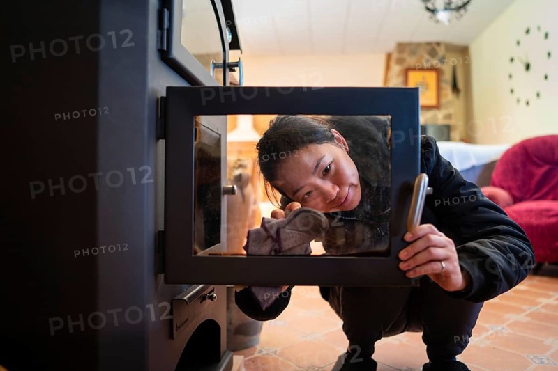 An Asian woman is cleaning the glass door of a wood burning stove, ensuring it is spotless for efficient use. She is indoors, with a cozy home background featuring warm colors