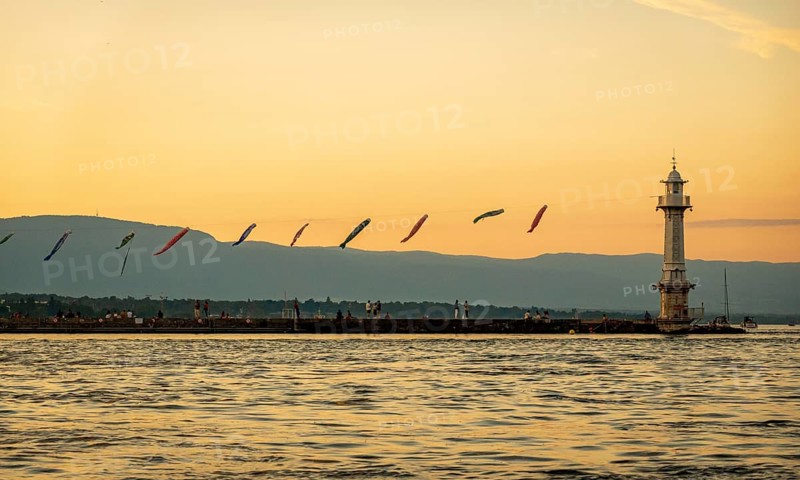 A serene sunset bathes Lake Geneva (Lac Leman) in warm hues, highlighting a lighthouse and vibrant kites in the sky, with the silhouette of distant mountains