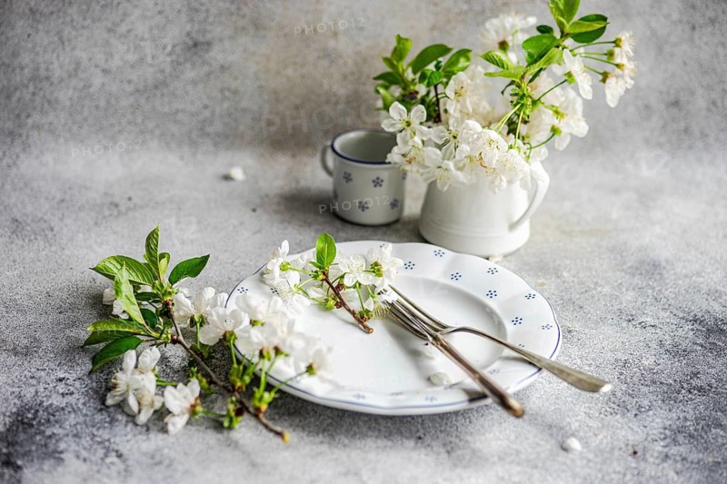 An elegant dining scene featuring a white plate adorned with delicate flowers, a fork, and a knife. A white jug with matching blossoms complements the aesthetic