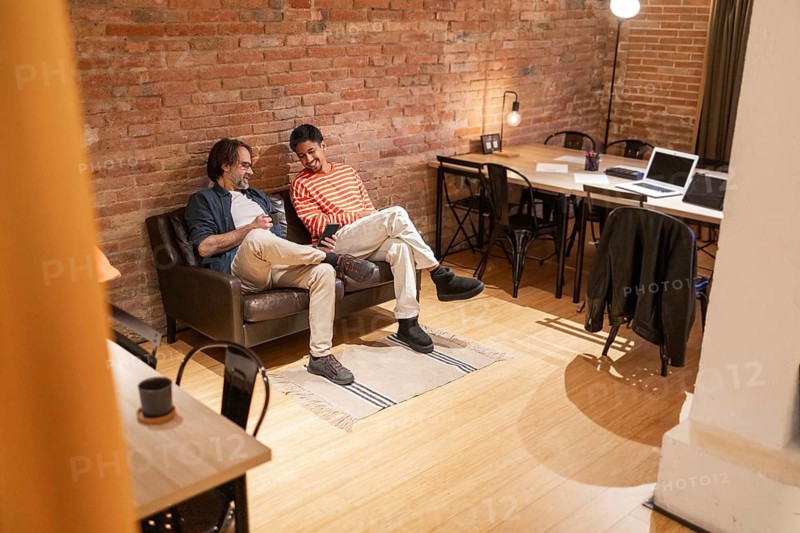 Two men sit on a sofa in a modern coworking space with brick walls and wooden floors. A laptop and table are in the background, fostering productivity and collaboration