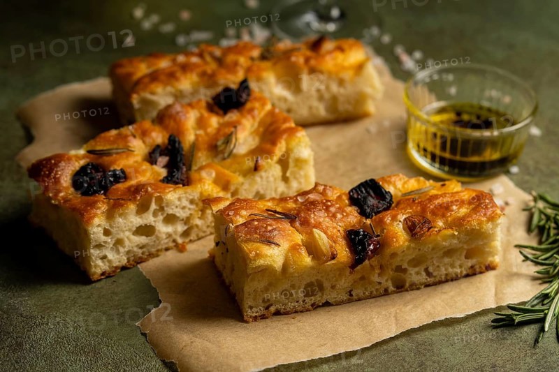 Close up of freshly baked homemade focaccia slices topped with rosemary and olives, served on parchment paper, accompanied by a small bowl of olive oil for dipping