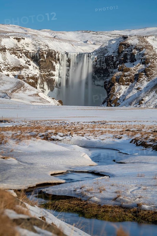 Skogafoss waterfall, surrounded by snow-covered terrain under a clear blue sky. The frozen landscape and flowing water create a stunning winter scene