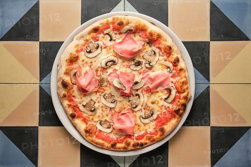 Round pizza with ham and mushrooms served on white plate on colorful tiled table