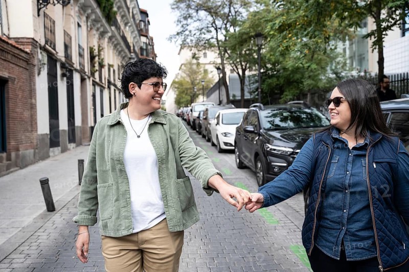 A curvy lesbian couple holds hands as they enjoy a stroll down a charming city street, surrounded by parked cars and trees. They share a look of happiness and connection