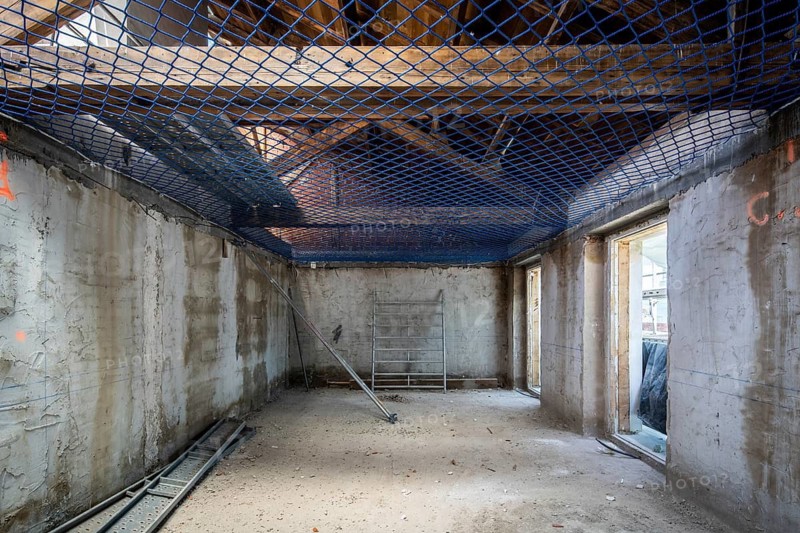 Interior view of a construction site featuring concrete walls, exposed beams, and safety netting. Scaffolding is set up for ongoing work. Natural light filters through window openings