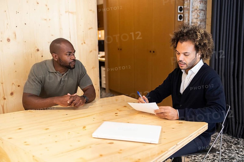 Two men engage in a focused meeting in a coworking space in Barcelona, seated at a modern wooden table, fostering collaboration and creativity within a professional environment