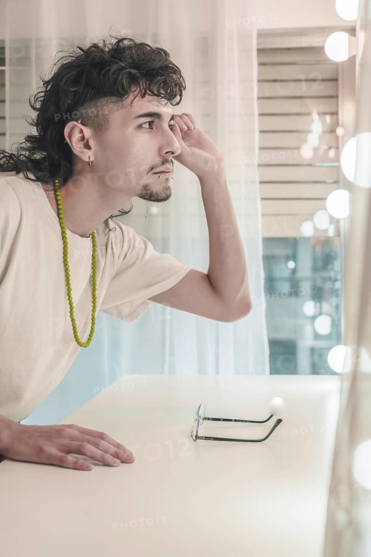 Young artist with a mullet and goatee, wearing a white t-shirt and green beaded necklace, admiring his reflection in a lighted mirror while touching his face