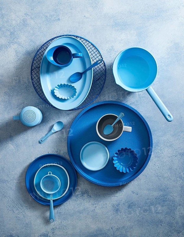 Top view of a stylish blue ceramic crockery set arranged on a textured gray background. The set includes plates, bowls, cups, and utensils, all showcasing a vibrant cobalt blue hue