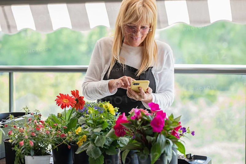 A gardener in an apron uses a smartphone, surrounded by vibrant potted flowers, on a balcony. The scene blends technology with nature in a peaceful setting