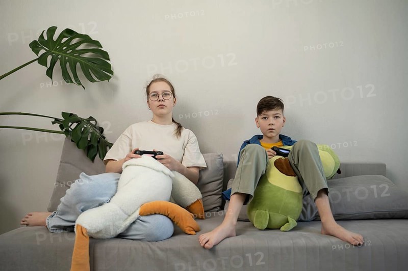 Two kids sit on a couch, engrossed in video gaming. Each holds a controller and is surrounded by plush toys, exuding a relaxed and focused atmosphere