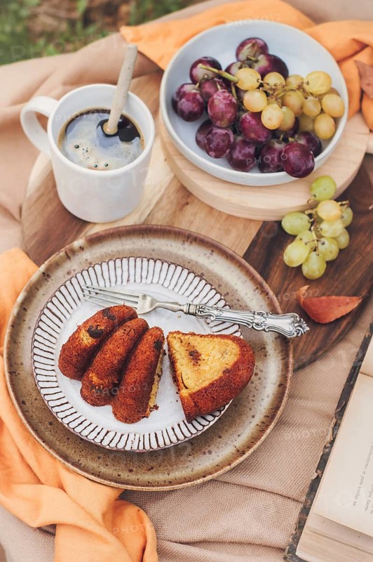 A slice of pumpkin cake is artfully plated with a fork on a rustic dish, accompanied by a cup of coffee and a bowl of grapes, creating a cozy autumn setting