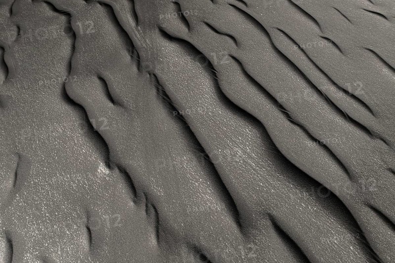 A detailed view of undulating black sand patterns left naturally sculpted by wind and water, highlighting the unique texture and reflective sparkles in sunlight